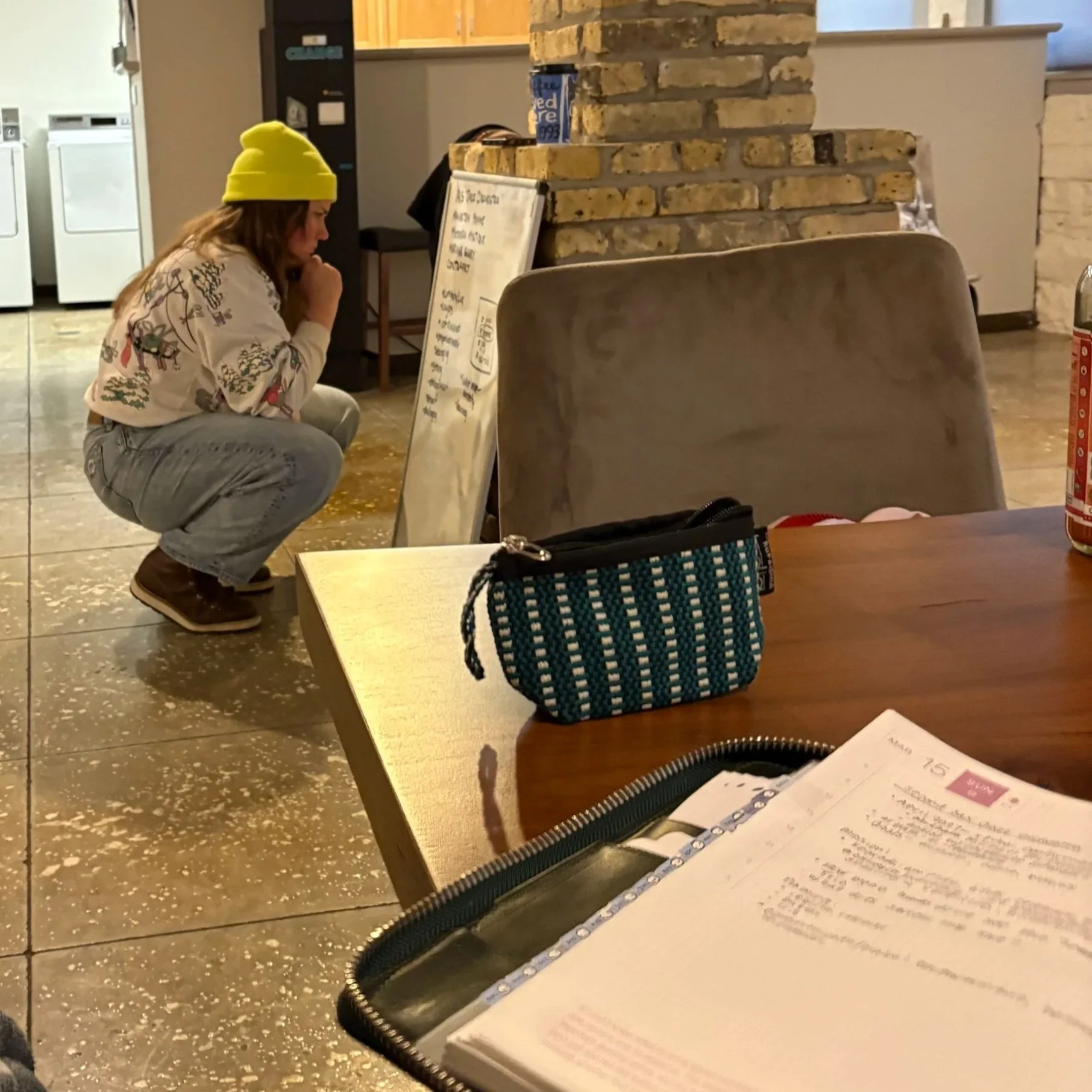 A woman in a yellow beanie, squatting on a stepping stone, looking at a sign or menu board in a cozy indoor setting, possibly a cafe or restaurant, with other furniture and objects around.