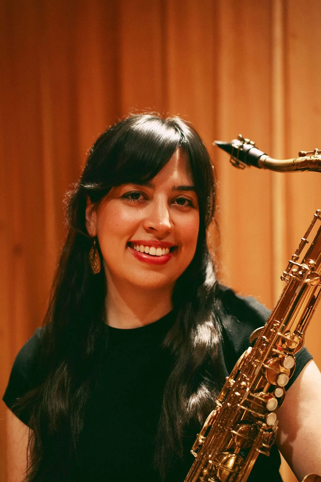 A woman with long dark hair holding a saxophone and smiling, set against a wooden background.