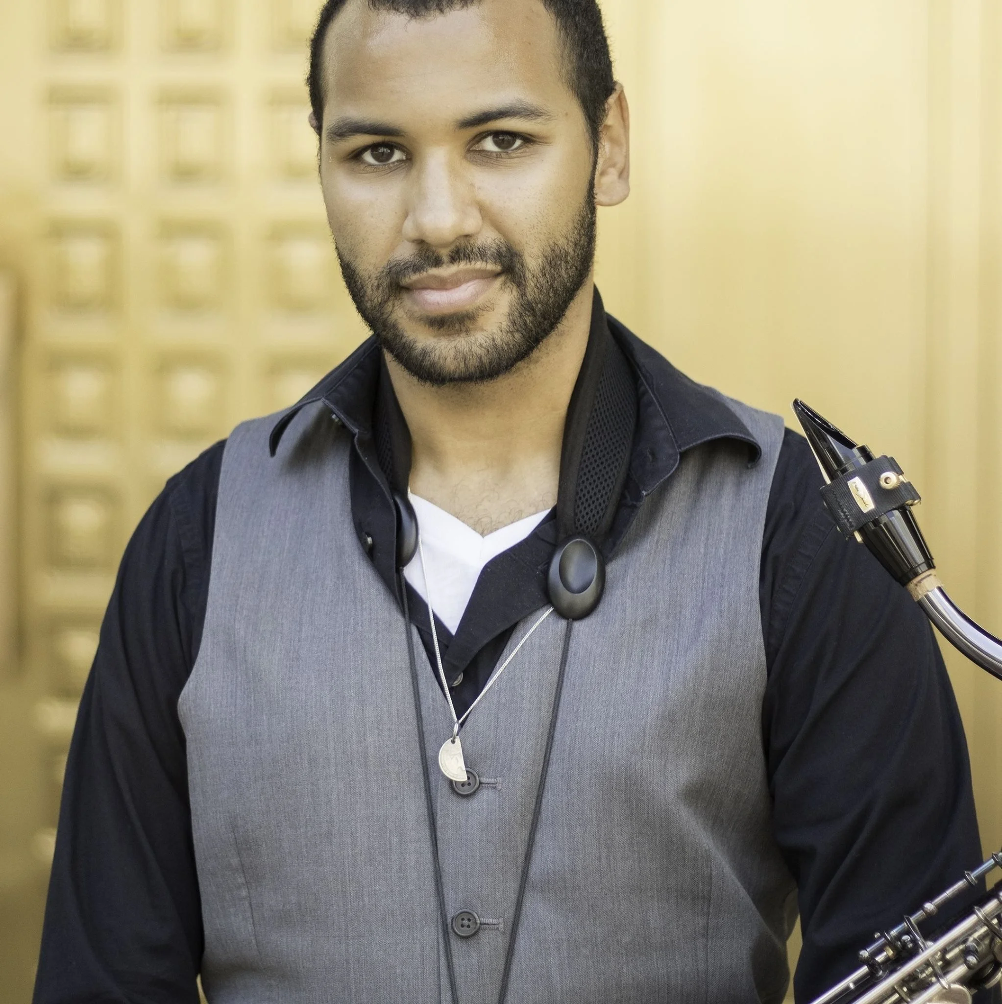 A man with short curly hair, a beard, and a mustache holding a trumpet, wearing a dark shirt and a gray vest, standing in front of a yellow background.
