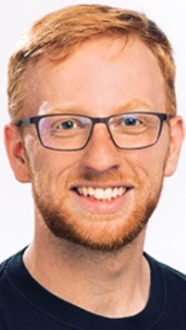 A smiling man with ginger hair, glasses, and a beard, wearing a dark shirt.