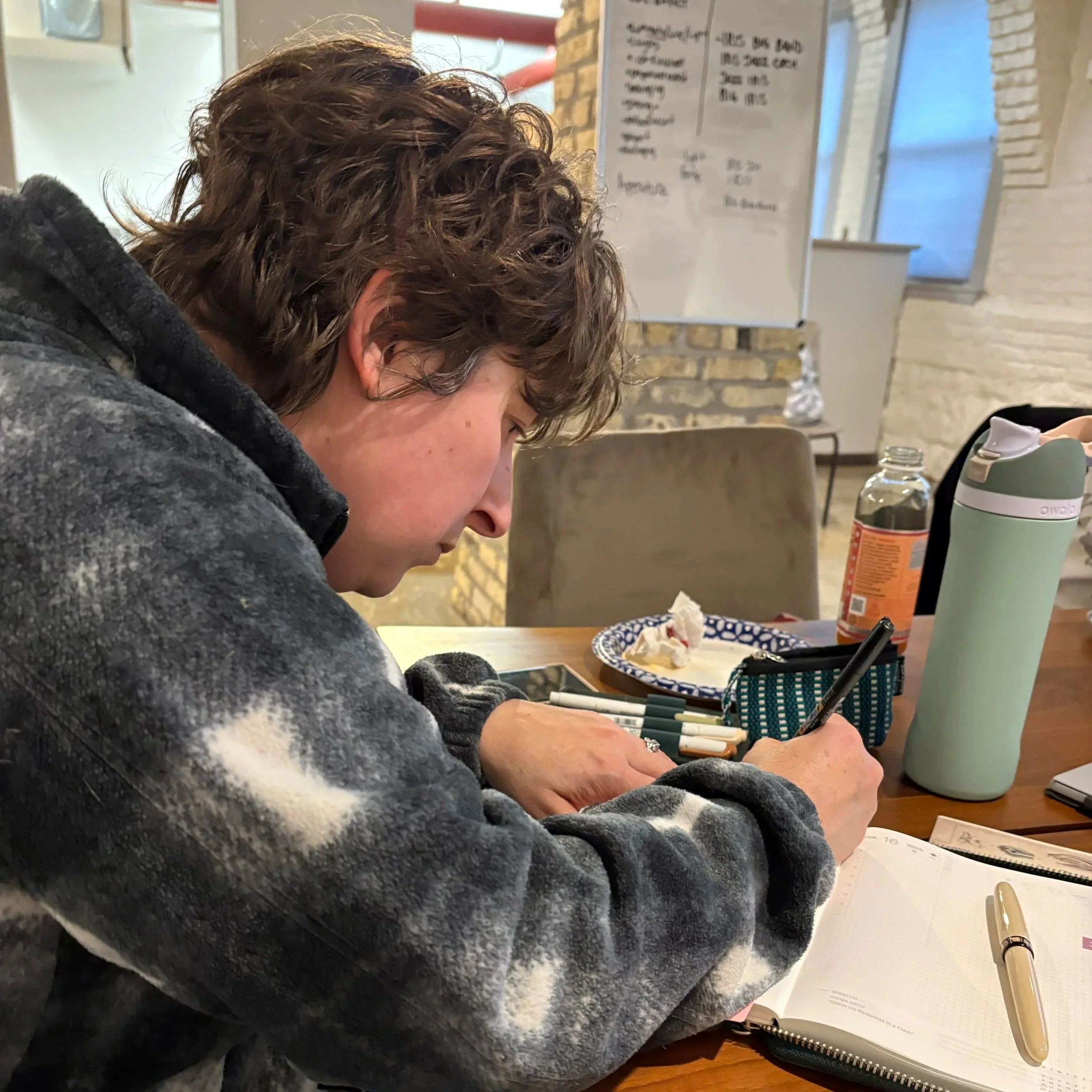 A person with curly brown hair and wearing a gray hoodie is sitting at a table, looking at a notebook. On the table, there are reading glasses, a water bottle, and some pens. In the background, there is a whiteboard with handwritten notes and a brick wall.