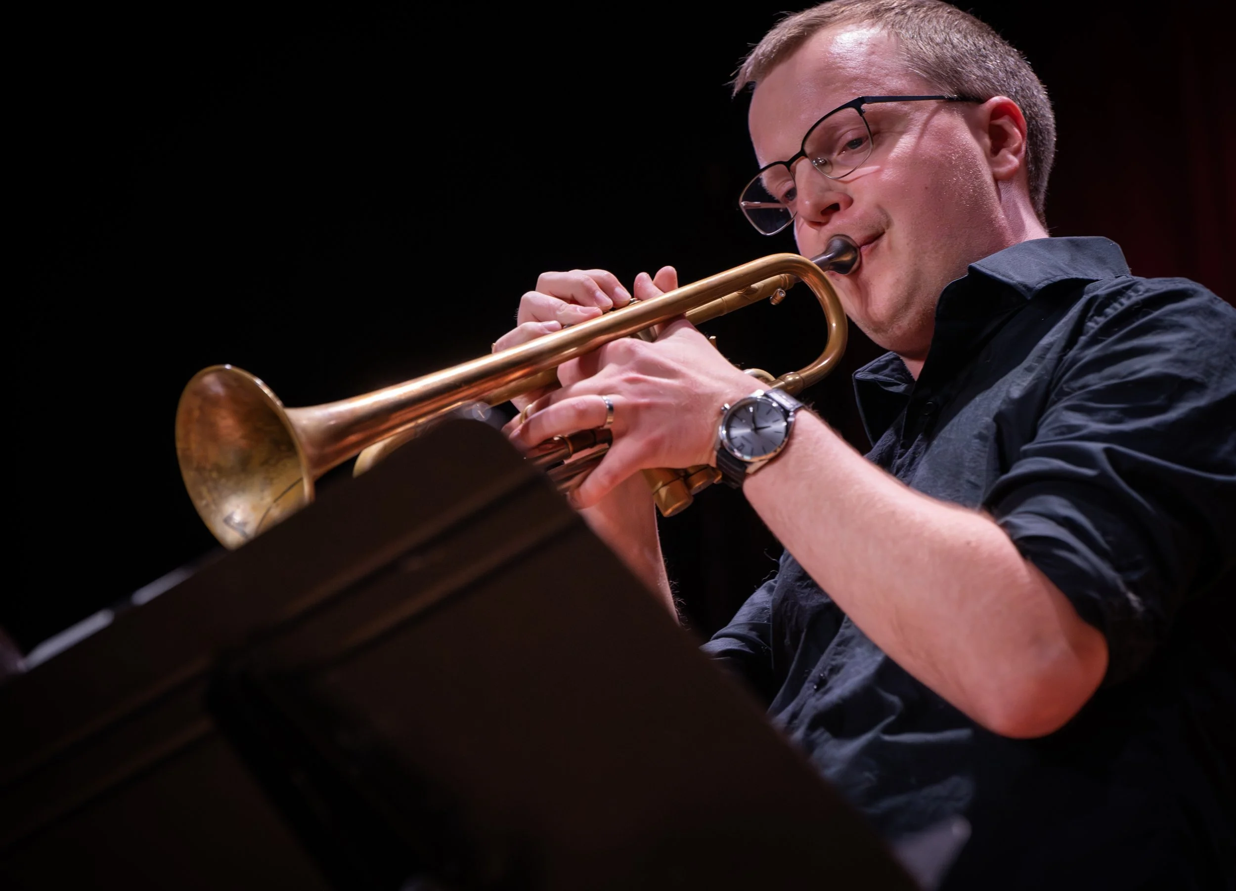 Musician wearing glasses plays a brass trumpet on stage.