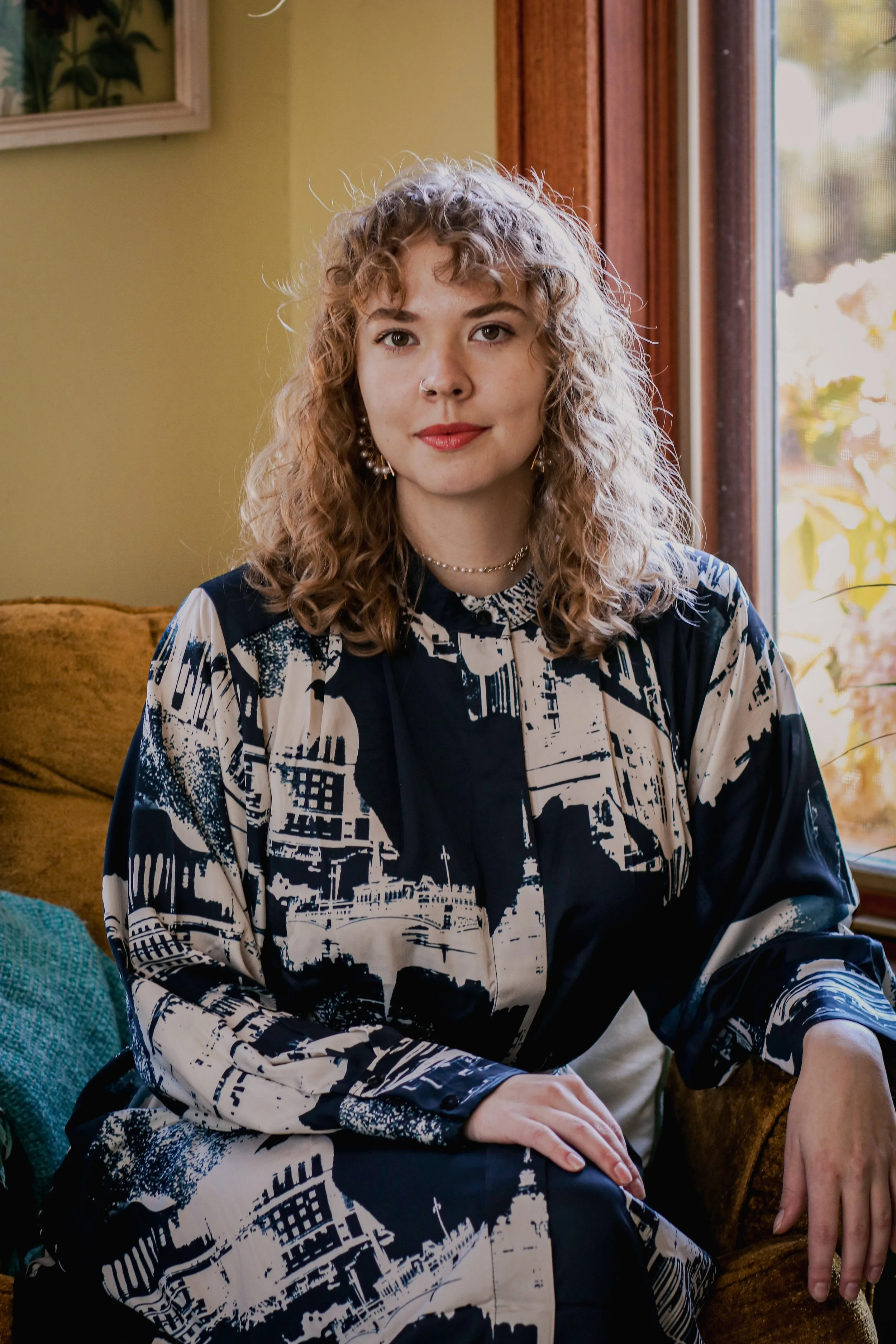 A young woman with curly blonde hair, wearing makeup and jewelry, sitting on a brown velvet couch near a window.