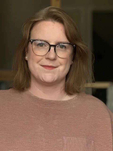 A woman with shoulder-length light brown hair, wearing glasses and a brown sweater, smiling at the camera.