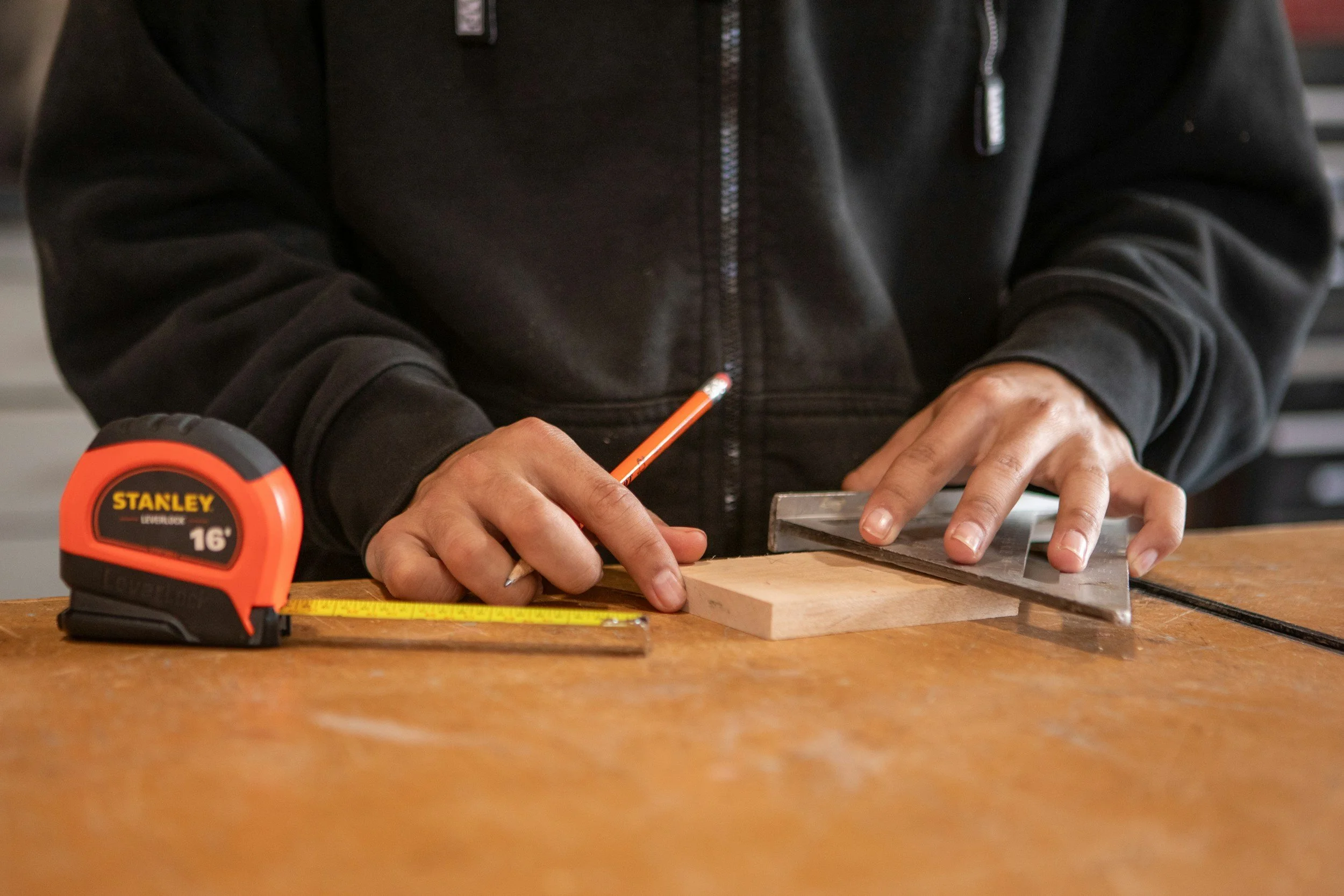 Person wearing a black hoodie measuring a piece of wood on a workbench, with a tape measure and a pencil nearby.
