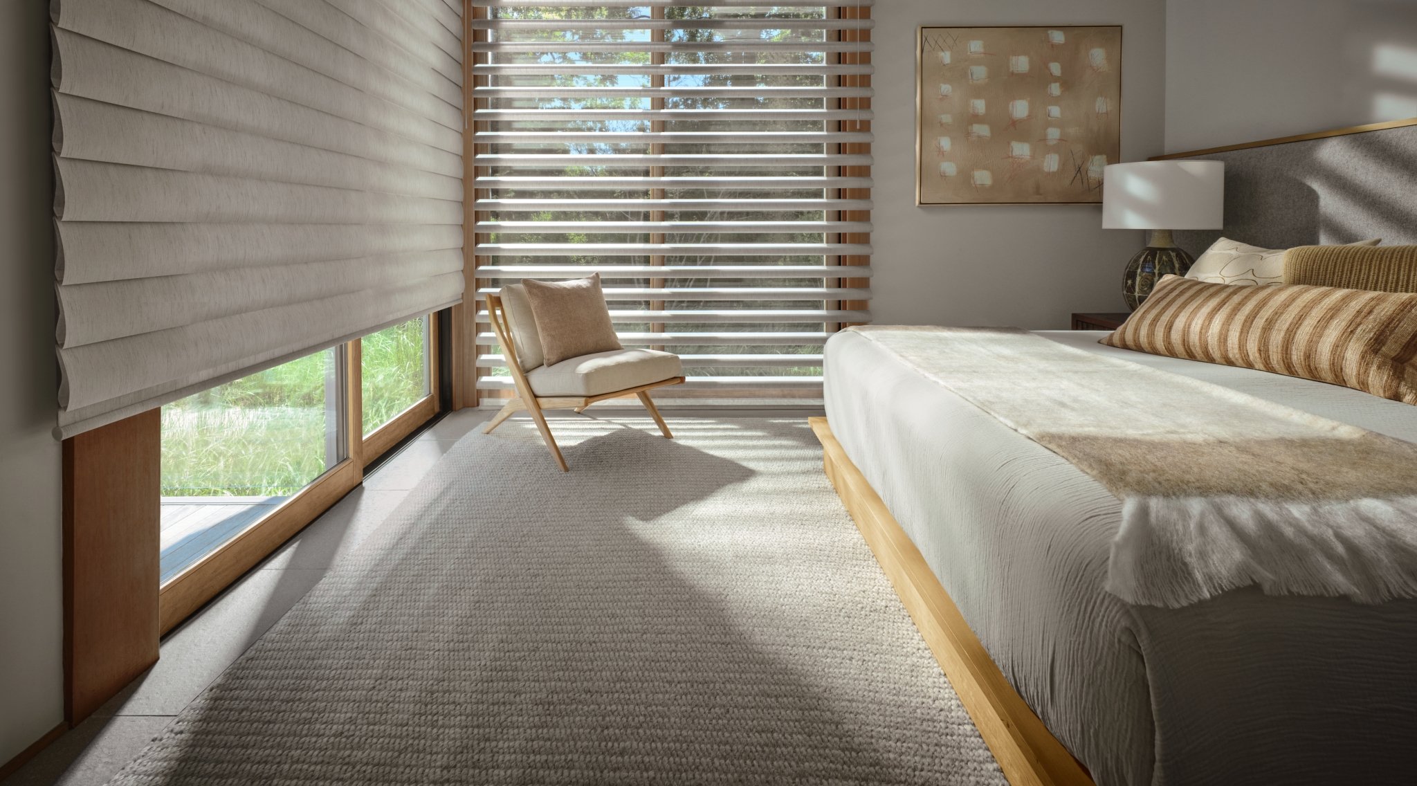 A cozy modern bedroom with large windows, Pirouette window shades, a bed with striped pillows, a side table with a lamp, a chair with a cushion, and abstract wall art.