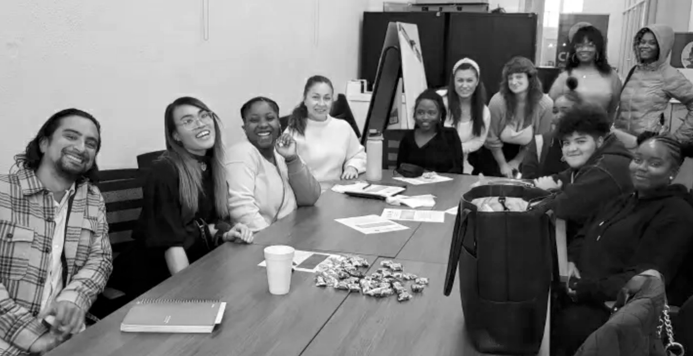 Group of twelve diverse young adults sitting and standing around a conference table in a meeting room, smiling and looking at the camera.