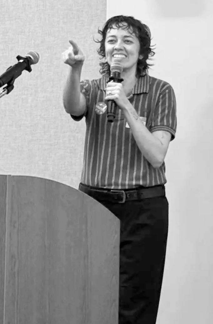 A person with short curly hair speaking into a microphone, standing behind a podium, gesturing with her right hand, smiling in a striped shirt.