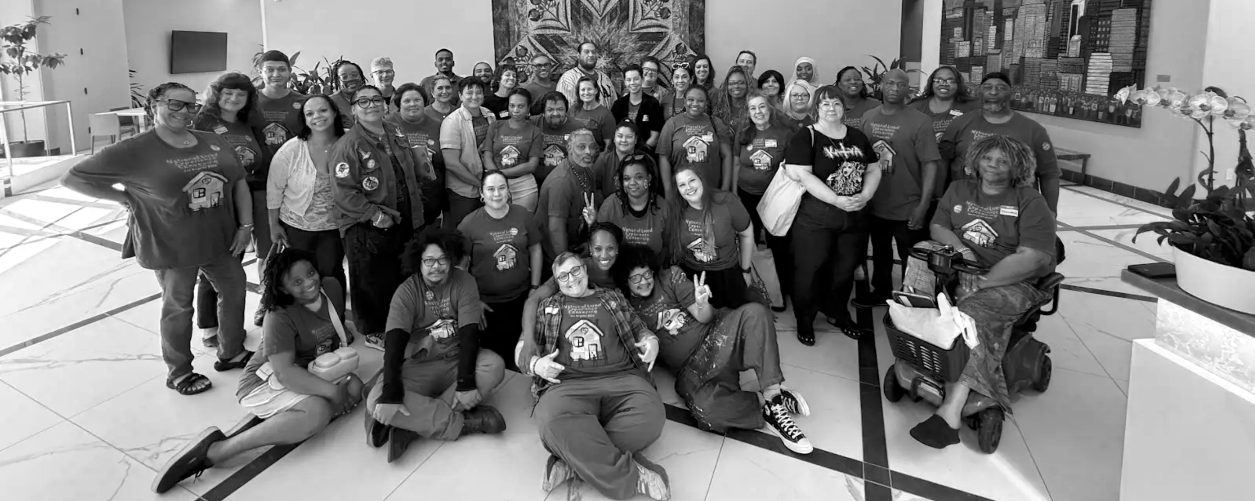 Group photo of diverse people gathered indoors, many wearing matching T-shirts with a house logo, some smiling, some making peace signs, in a lobby or common area with artwork and plants.