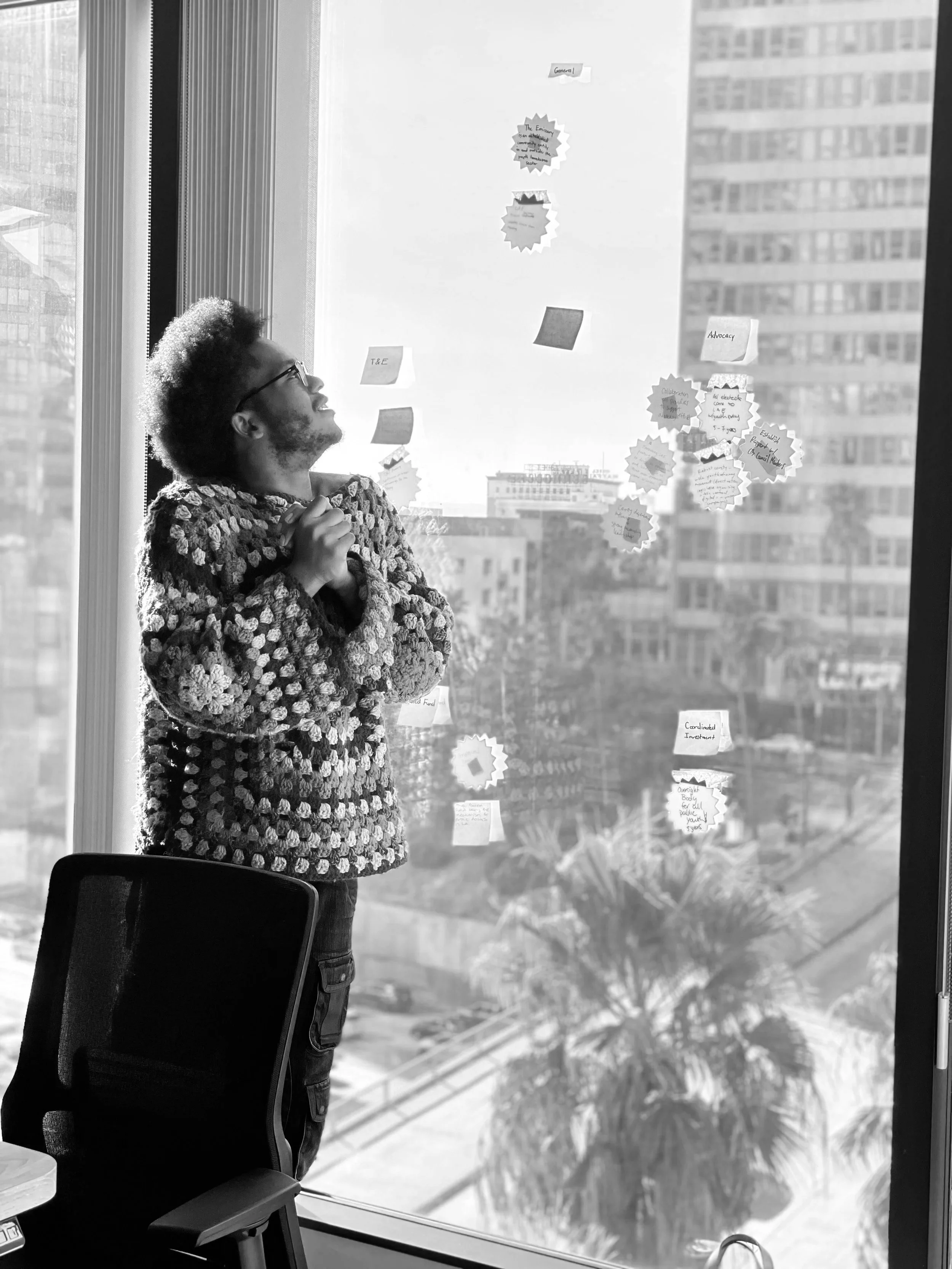 A man with glasses and curly hair looking up at a wall of sticky notes and papers on a glass window, with a cityscape visible outside.