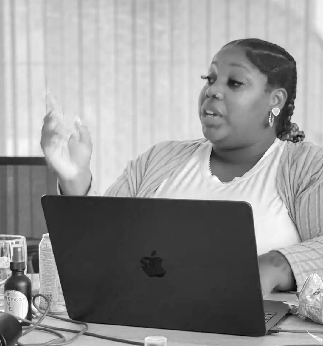 A woman sitting at a table with a laptop, gesturing with her right hand, during a discussion or presentation.