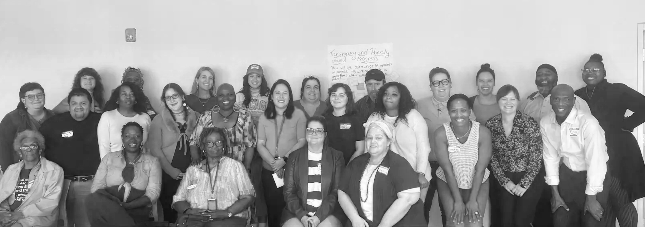 Group of diverse people smiling for a photo indoors, some wearing name tags, in front of a wall with a poster about transparency and honesty.