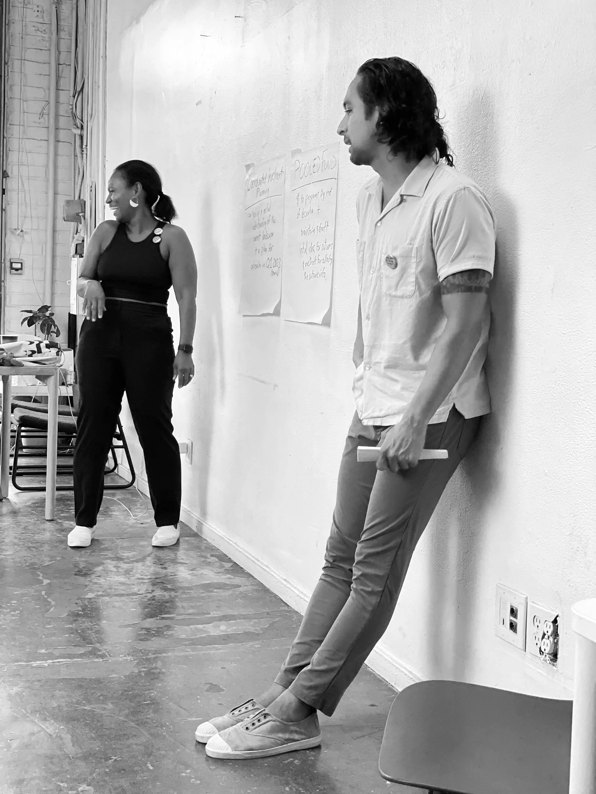 Two people stand and lean against a wall in a room with a whiteboard.