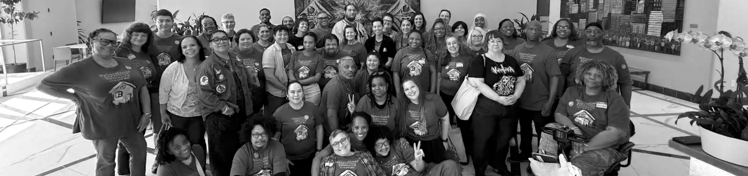 A large group of diverse people gathered indoors for a photo, some smiling and making peace signs, with plants and artwork in the background.