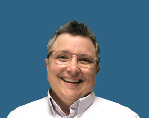 A smiling person with short gray hair, glasses, and a white collared shirt against a blue background.