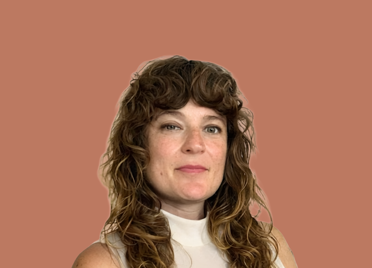 A woman with curly brown hair and blue eyes, wearing a white top, standing against a plain brown background.