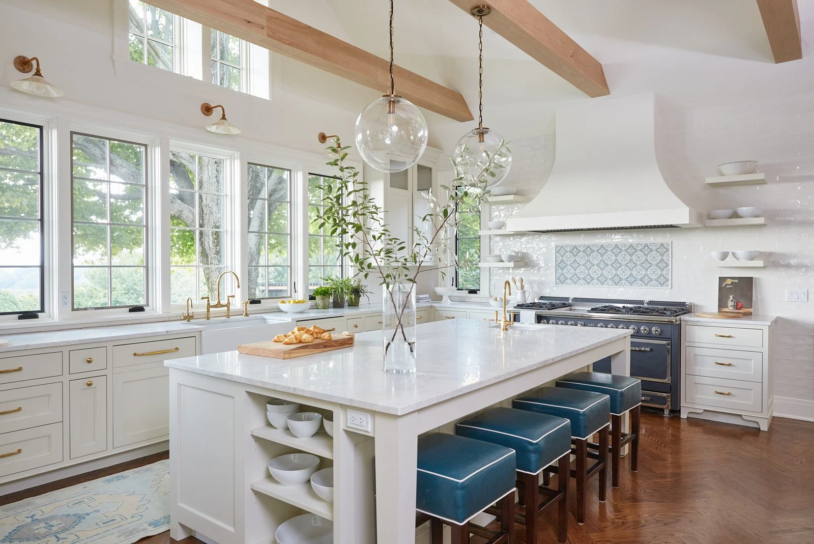 Bright white kitchen with large windows, white cabinets with gold hardware, a central island with a vase of branches, four blue barstools, and a professional-grade stove with a decorative tile backsplash.