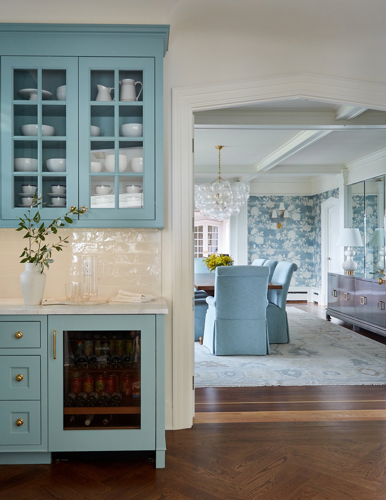 A view into a dining room from a kitchen area, showing a blue china cabinet with white dishes and a wine refrigerator below, with a vase of greenery on the counter. The dining room has light blue upholstered chairs around a wood table, wallpaper with a floral pattern, and a chandelier. The flooring is dark hardwood with an area rug under the table.
