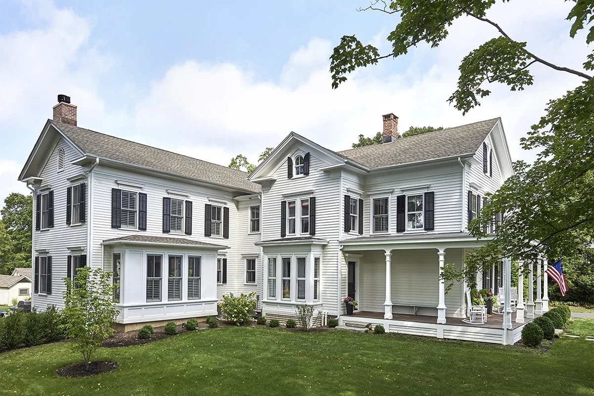 A large white two-story house with black shutters, a wrap-around porch with white columns, and a well-maintained green lawn with small bushes and trees.