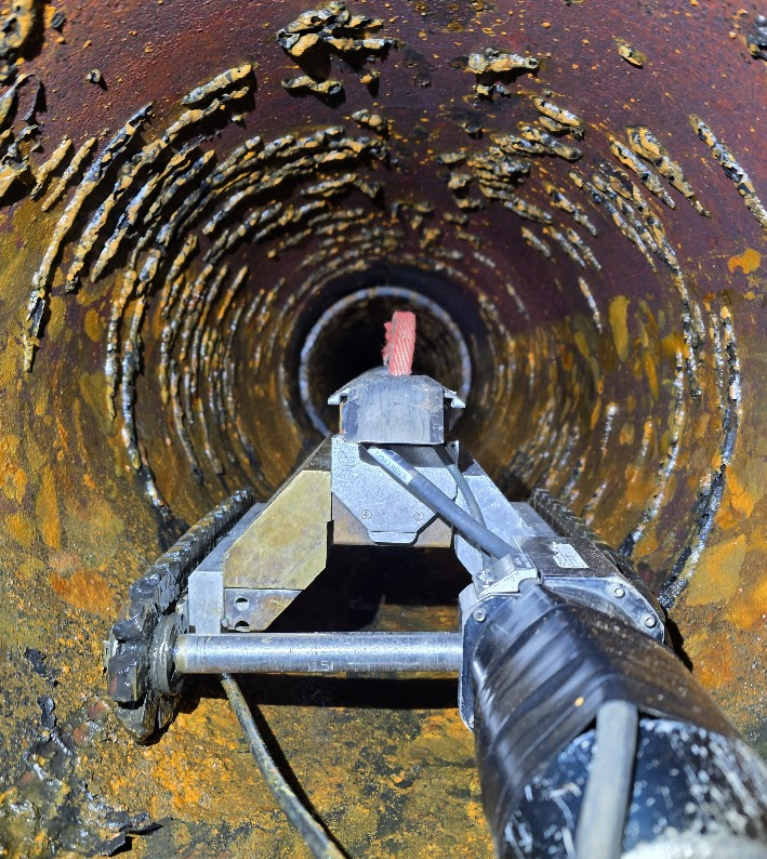 Inside a large, rusty pipe with a spiral pattern, a robot with a mechanical arm is inspecting the interior surface.