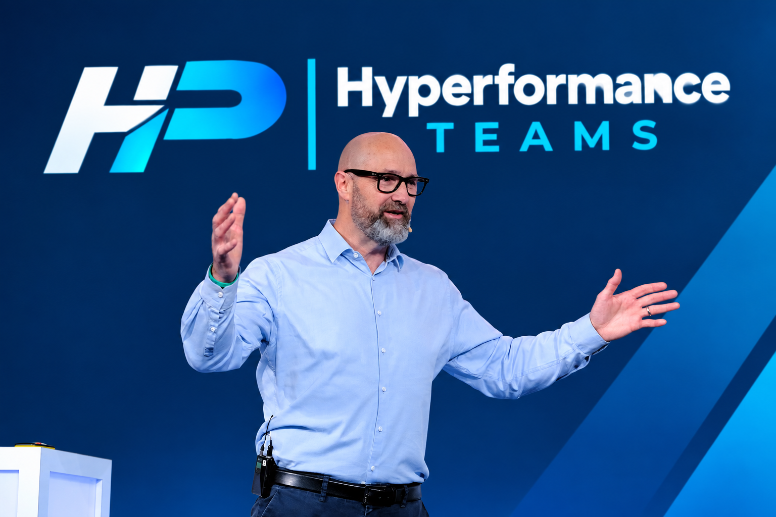 A man with glasses and a beard speaking at a conference in front of a blue background with 'Hyperformance Teams' and 'FP' logo.
