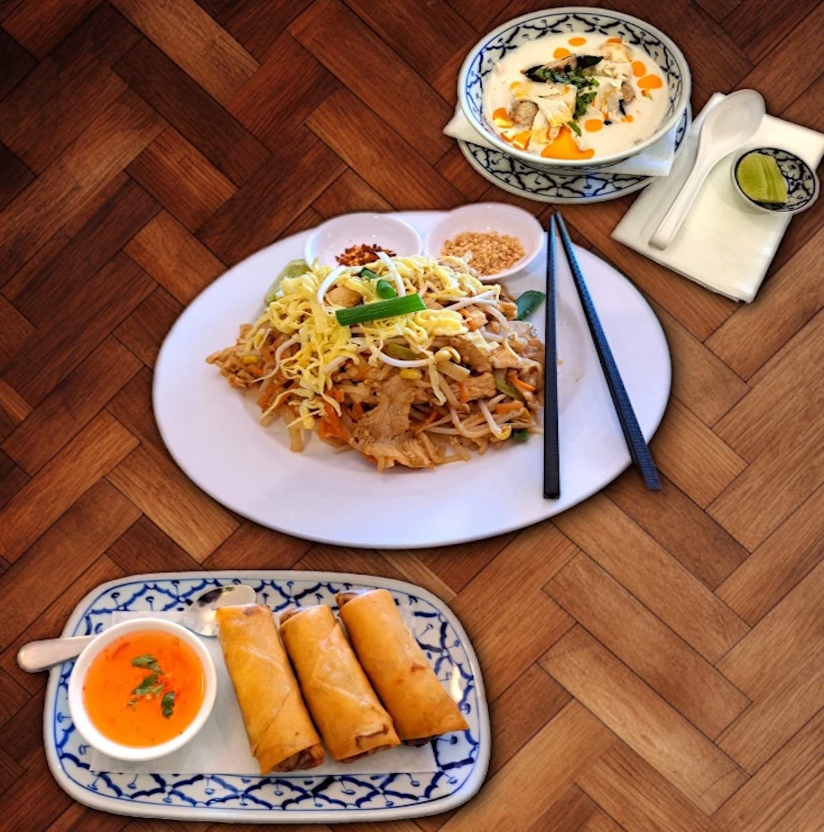 A meal featuring a plate of stir-fried noodles with vegetables and shredded egg, a bowl of Thai chicken coconut soup, and a plate of three spring rolls with dipping sauce, all on a wooden table.