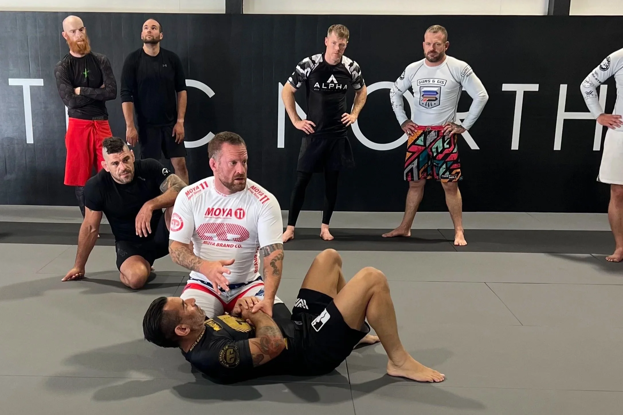 A group of men in athletic clothing training in a martial arts gym. Some are standing with hands on hips, others are practicing grappling or lying on mats.