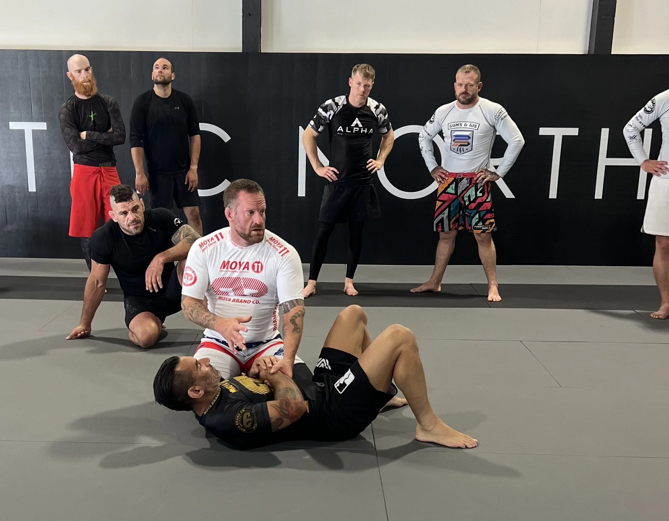 Group of martial artists practicing Brazilian Jiu-Jitsu in Electric North Jiu Jitsu,  with some on the mat and others standing against a black wall with white lettering, watching professor Greg Anderson teach.