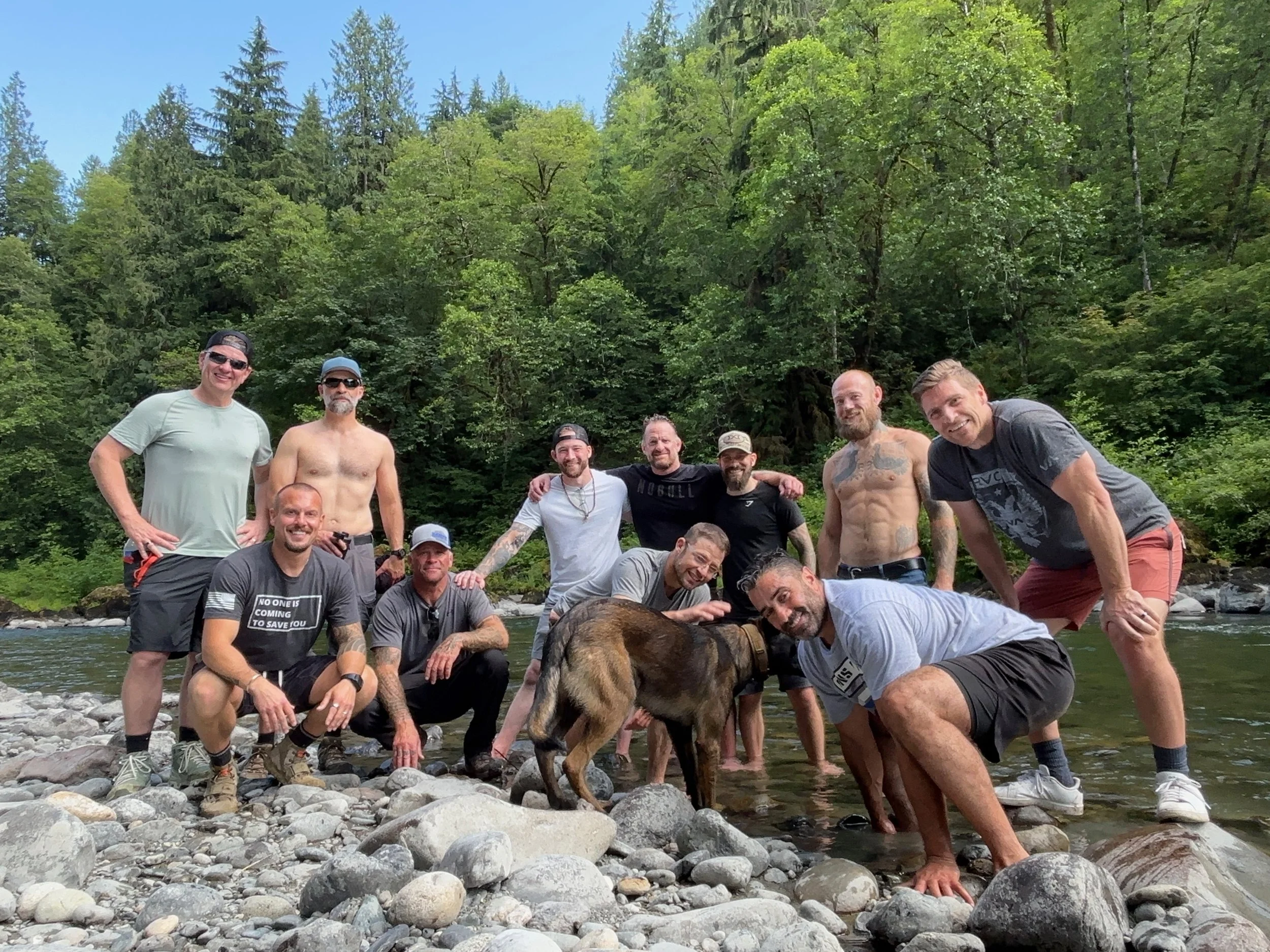A group of men posing in a river with large rocks, surrounded by lush green trees, with a dog in the foreground.