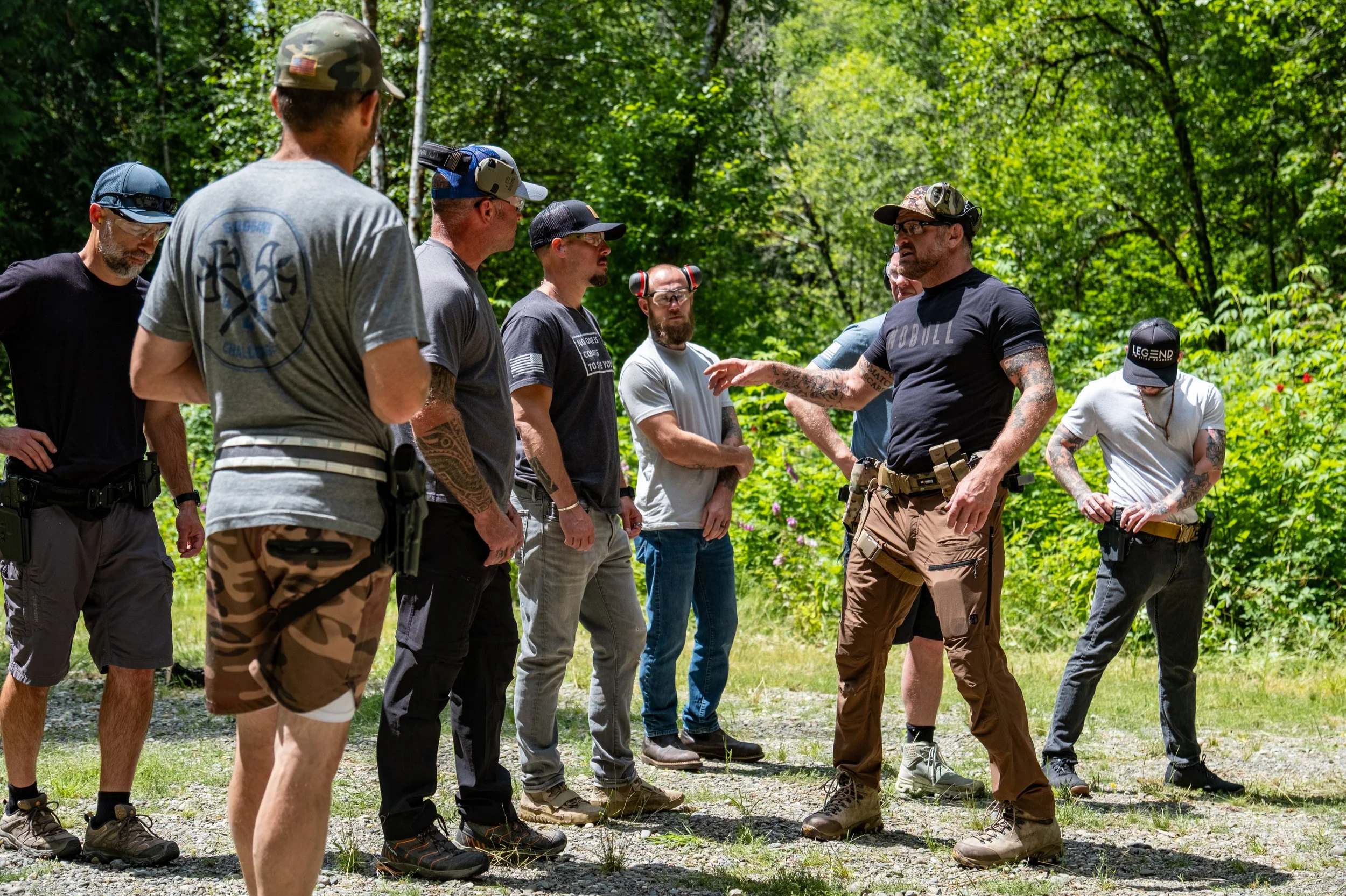 Group of men outdoors in a wooded area, engaged in a discussion, wearing casual and tactical clothing, some with hats and safety glasses.