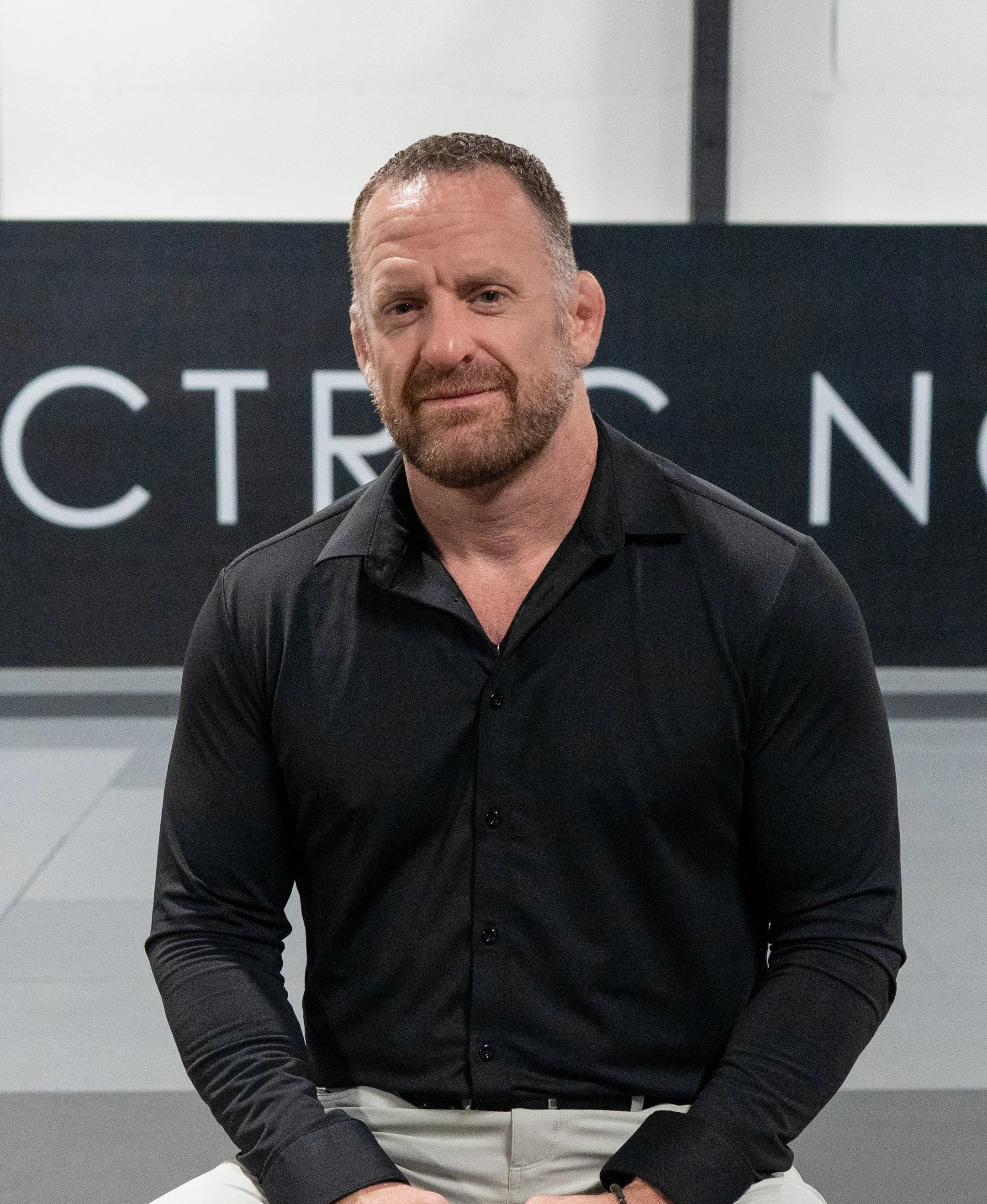 Greg Anderson, a man with a beard and short hair wearing a black shirt and beige pants, sitting against a modern black and white background in his Jiu Jitsu gym, Electric North Jit Jitsu.
