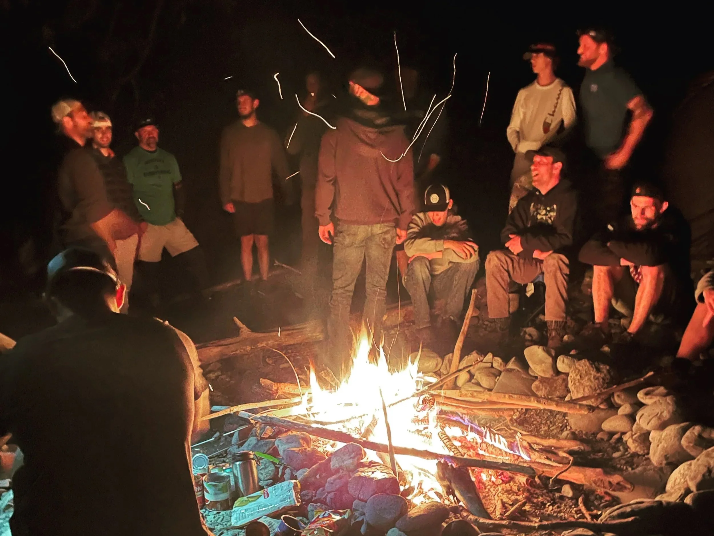 A group of people gathered around a campfire at night outdoors, with some standing and others sitting, enjoying the evening.