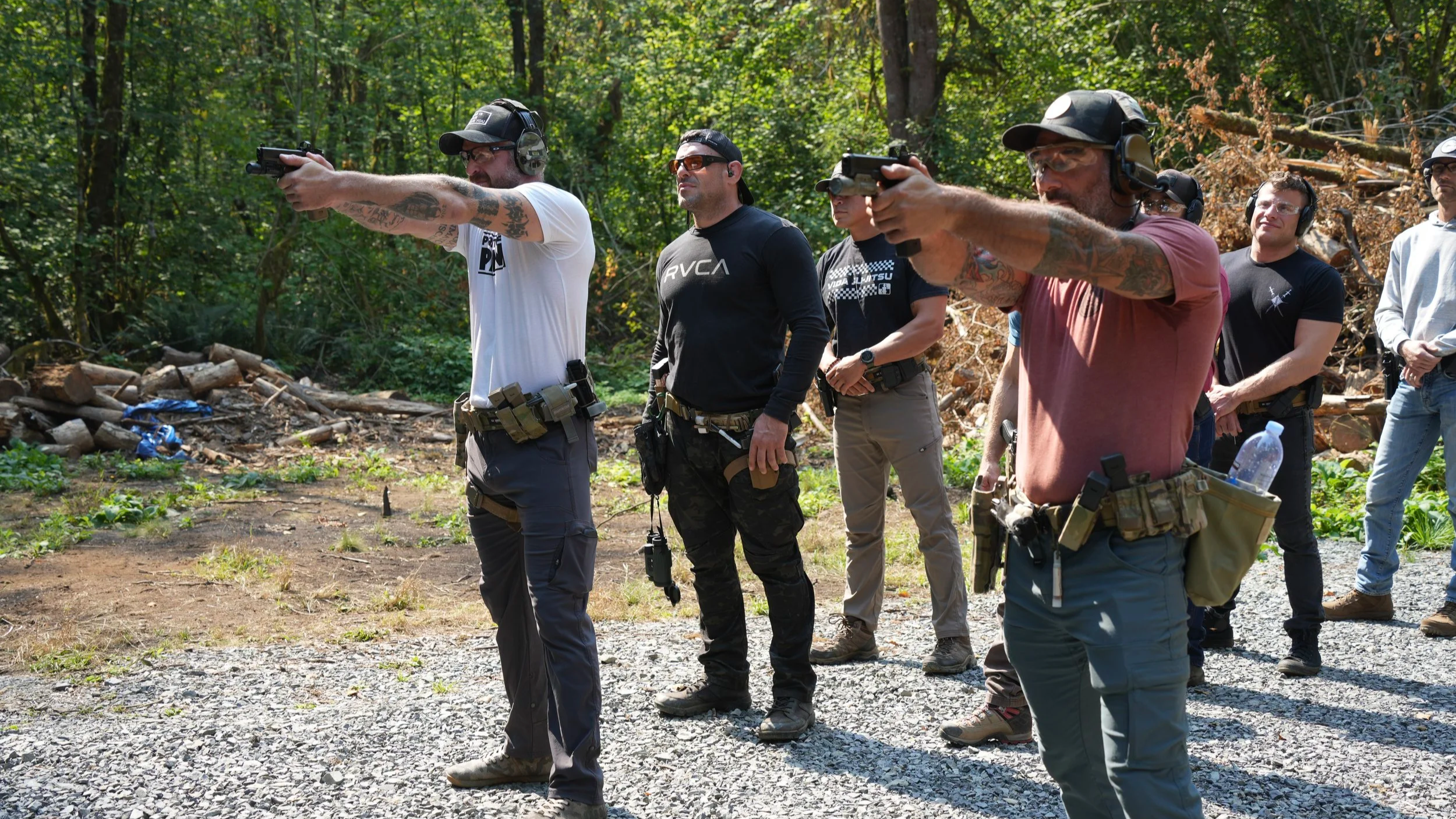 A group of men at a shooting range in a wooded outdoor setting, aiming pistols.
