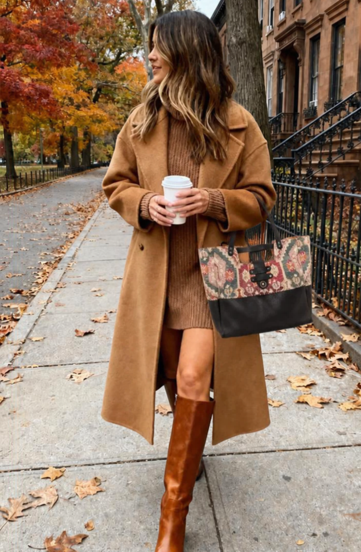 A woman dressed in a brown sweater, tan coat, boots, and carrying a floral patterned handbag is standing on a sidewalk by a tree with autumn leaves, holding a coffee cup and looking to her left.