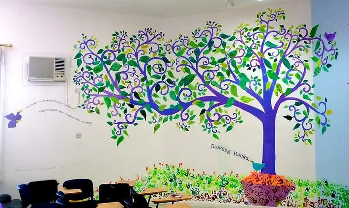 Colorful mural of a large purple and green tree with a hanging pot at the base, surrounded by green grass, with some purple and yellow birds and butterflies on a white wall in a room with chairs and an air conditioning unit.