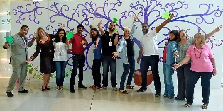 Group of ten diverse adults standing in front of a purple decorative wall mural, smiling, with some holding green party hats.