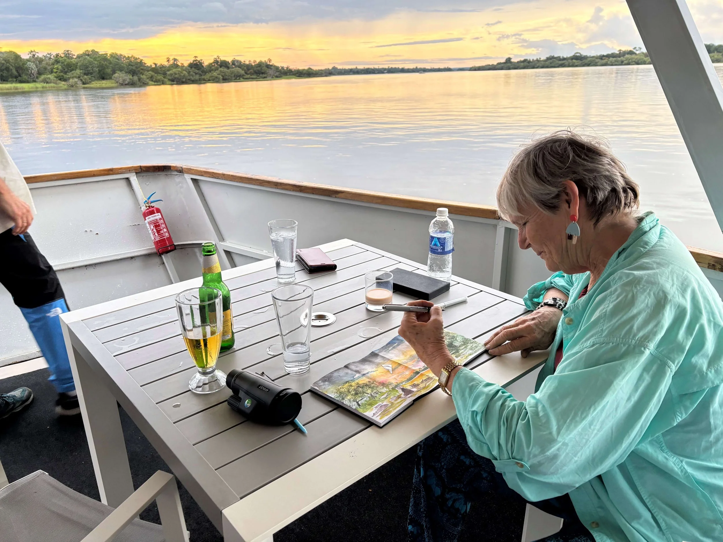 A woman is sitting at a white outdoor table on a boat, looking at a map. The table has various drinks, including a glass of beer, water bottles, and a glass of water. There is a telescope on the table. The boat is on a calm river with lush green tree
