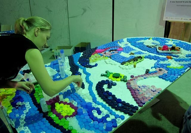 Person arranging colorful bottle caps on a large white surface to create a mosaic or artwork.