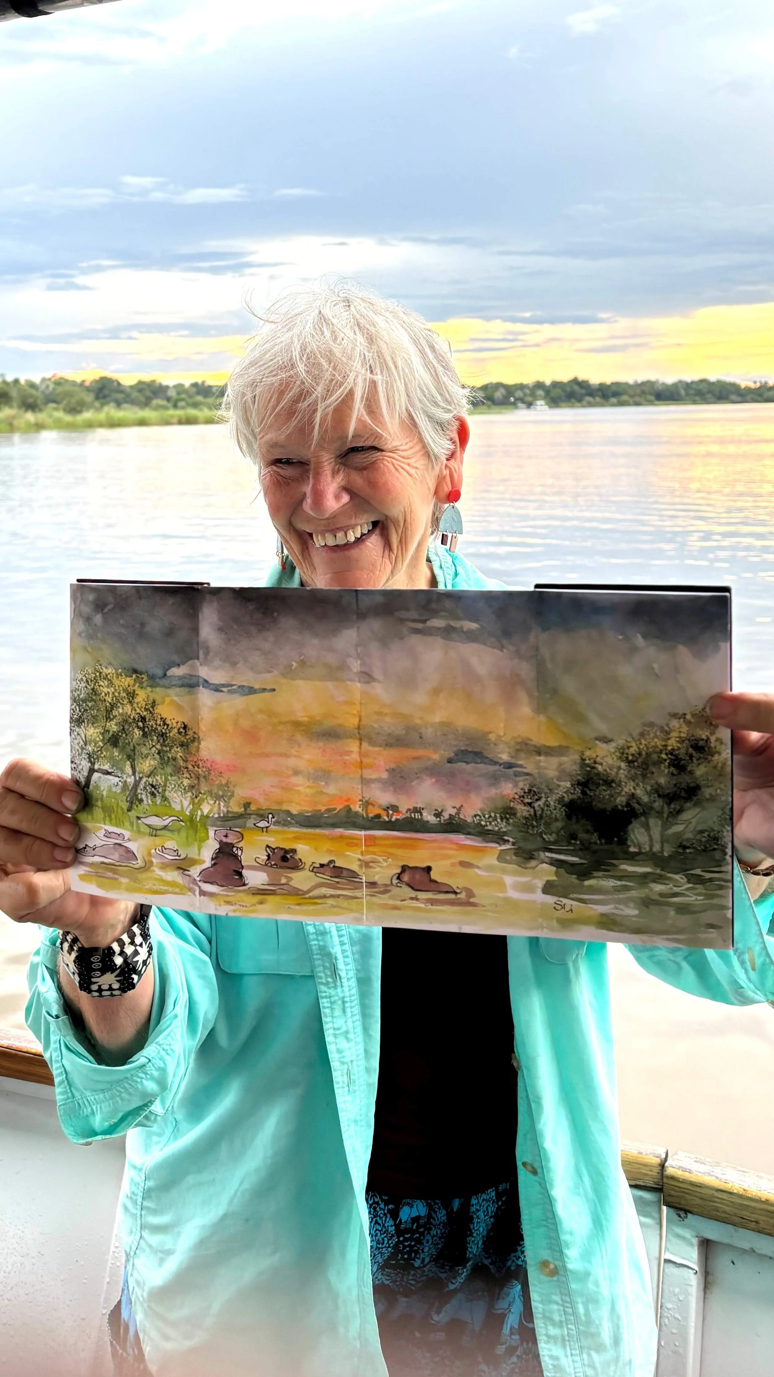 A woman with short gray hair and colorful earrings is holding an illustrated book with a landscape scene of a river, trees, and ducks at sunset. She is smiling and standing near a body of water with a cloudy sky overhead.