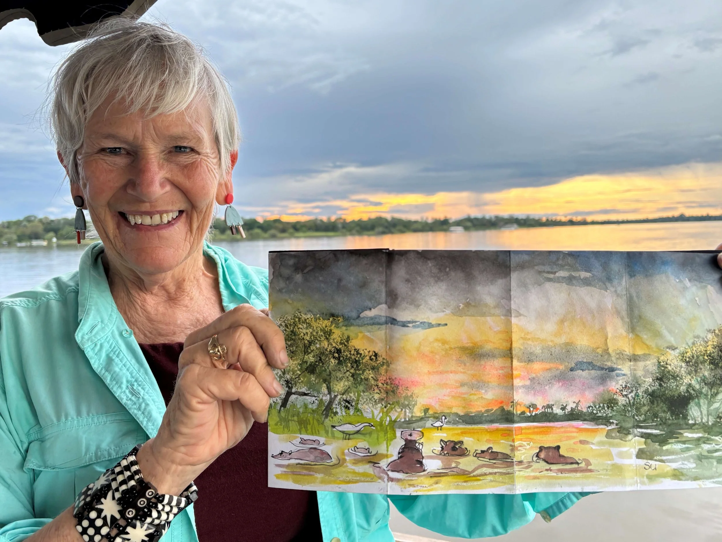 An older woman with short gray hair smiling and holding a colorful landscape painting outdoors near water during sunset. She is wearing a light turquoise jacket, earrings, and a black and white patterned bracelet.