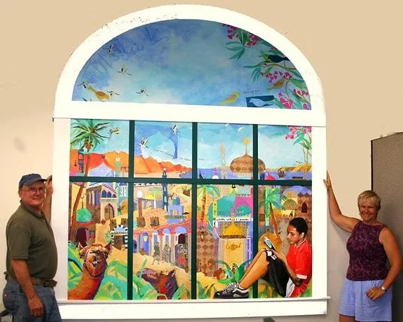 Two adults standing on either side of a large painted window with a vibrant cityscape featuring colorful buildings, palm trees, and a blue sky.