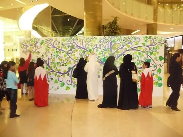 People in traditional Middle Eastern clothing, including abayas and thobes, standing and walking near a colorful painted wall in a shopping mall, with a group of people waiting in line on the left side.