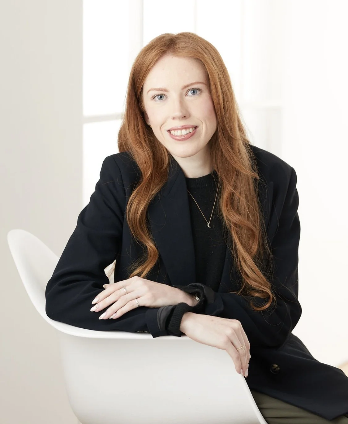 A young woman with long red hair and blue eyes, wearing a black blazer and necklace, sitting on a white chair in a bright room.