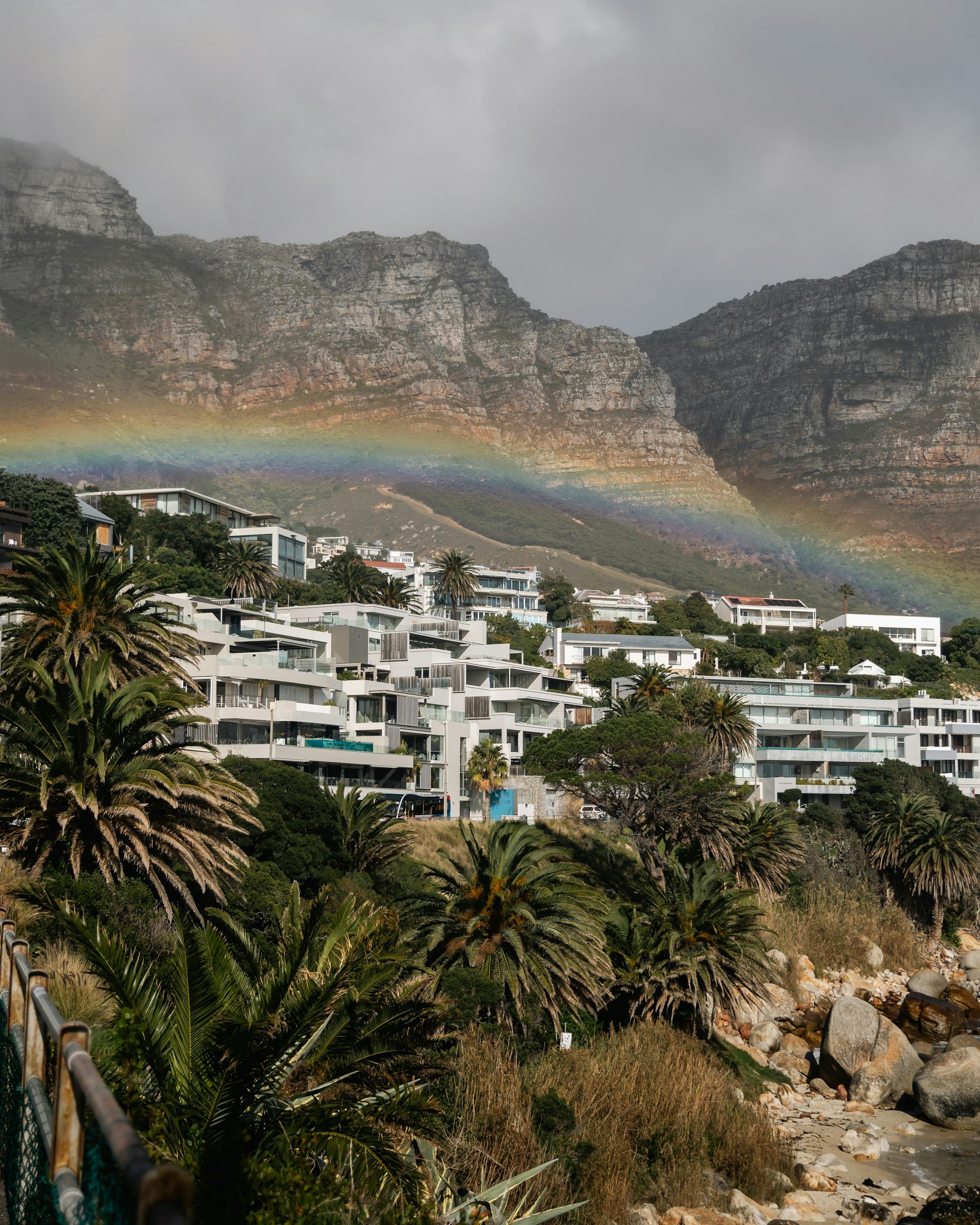 Residential buildings on a hillside with lush greenery, palm trees, and a rocky shoreline in the foreground. 12 Apostles in the background. A rainbow is faintly visible arching across the scene. This is the Atlantic Seaboard, Cape Town.