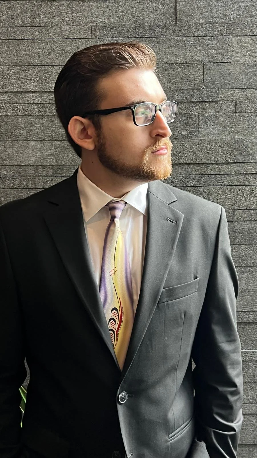 A young man with glasses, a beard, dressed in a black suit, white shirt, and a colorful tie, standing against a textured gray stone wall, looking to the side.