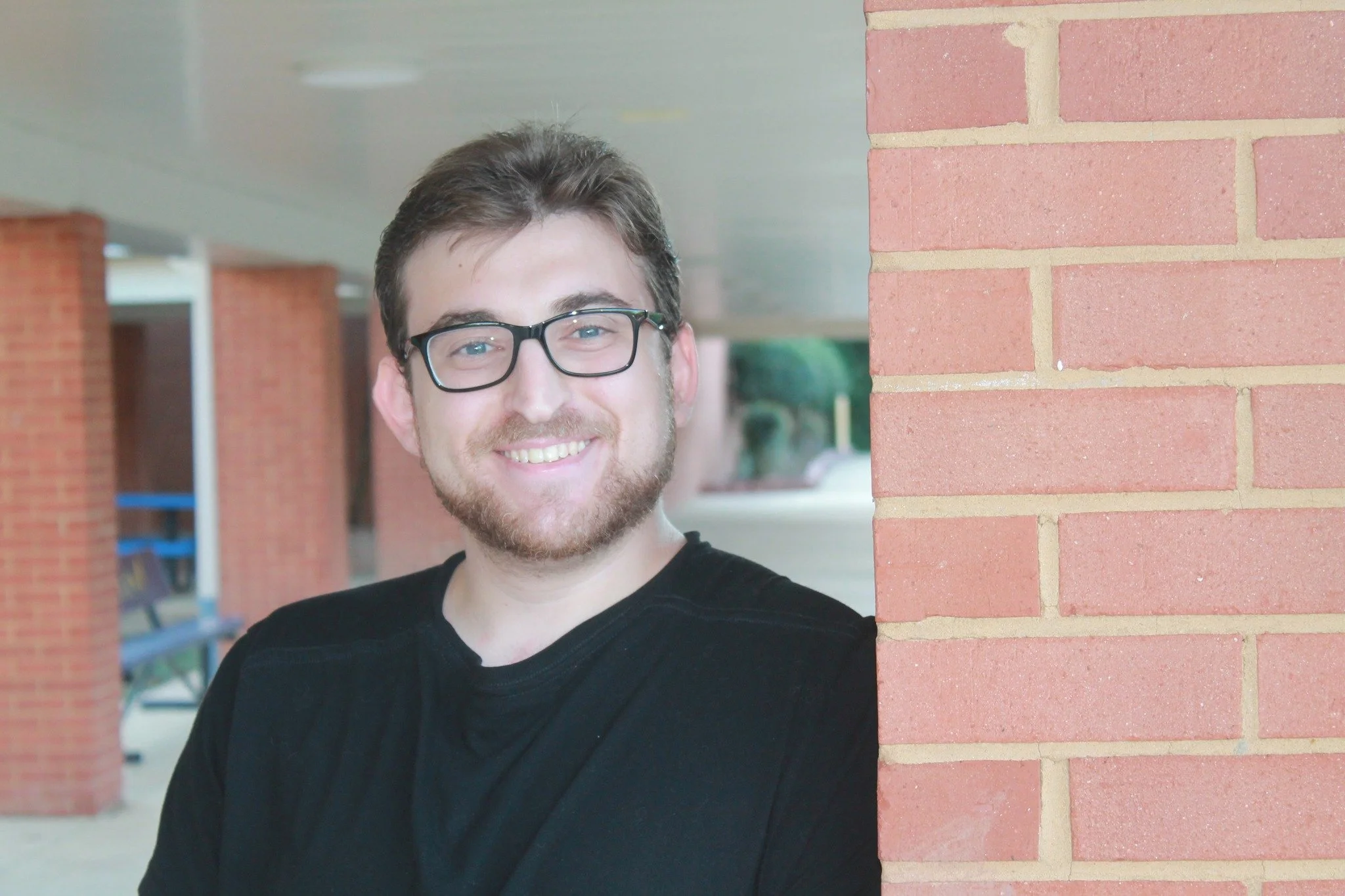 A young man with glasses and a beard smiling, standing next to a red brick wall in an indoor or semi-outdoor setting.