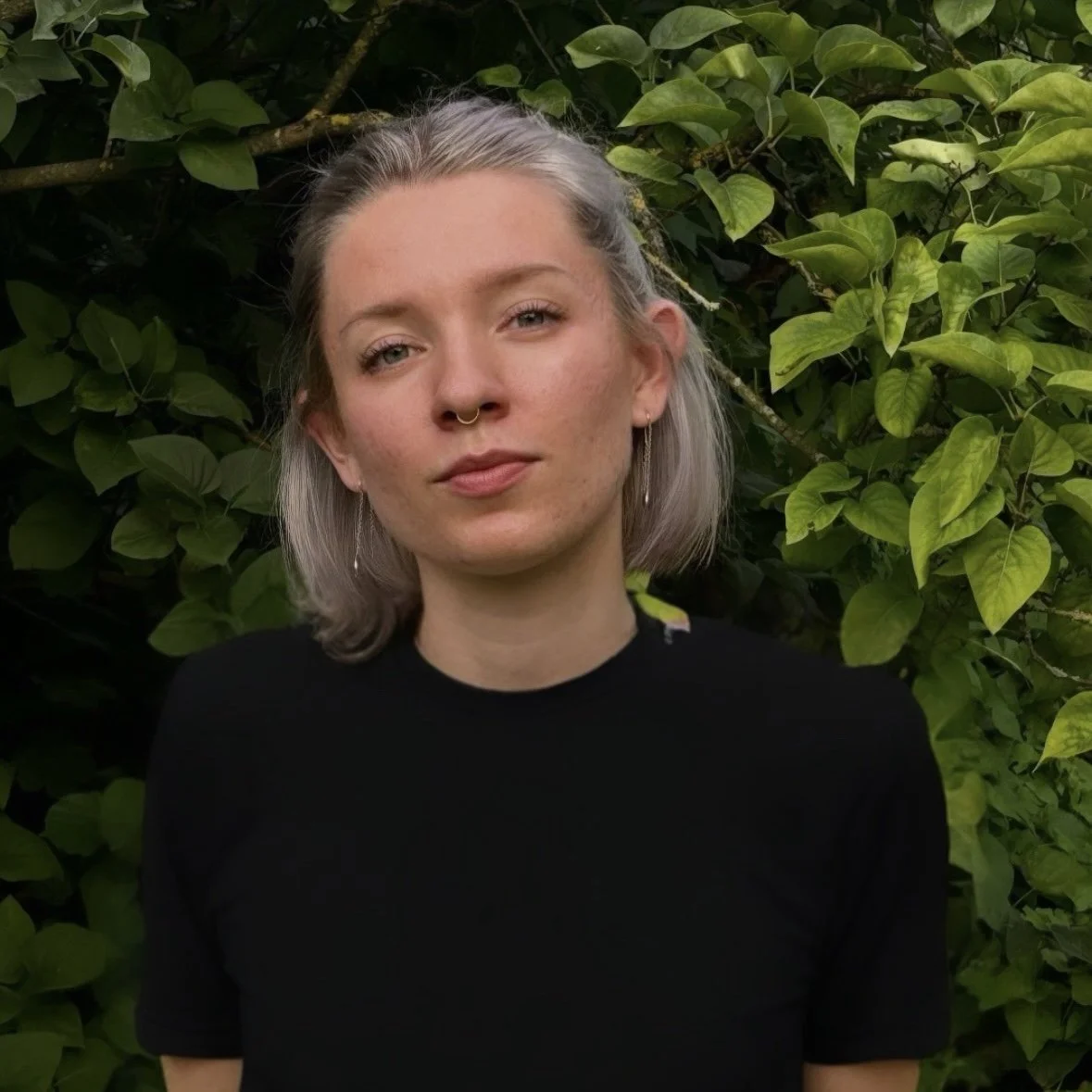 A young woman with light skin and short, grayish hair standing in front of dense green foliage, wearing a black shirt.