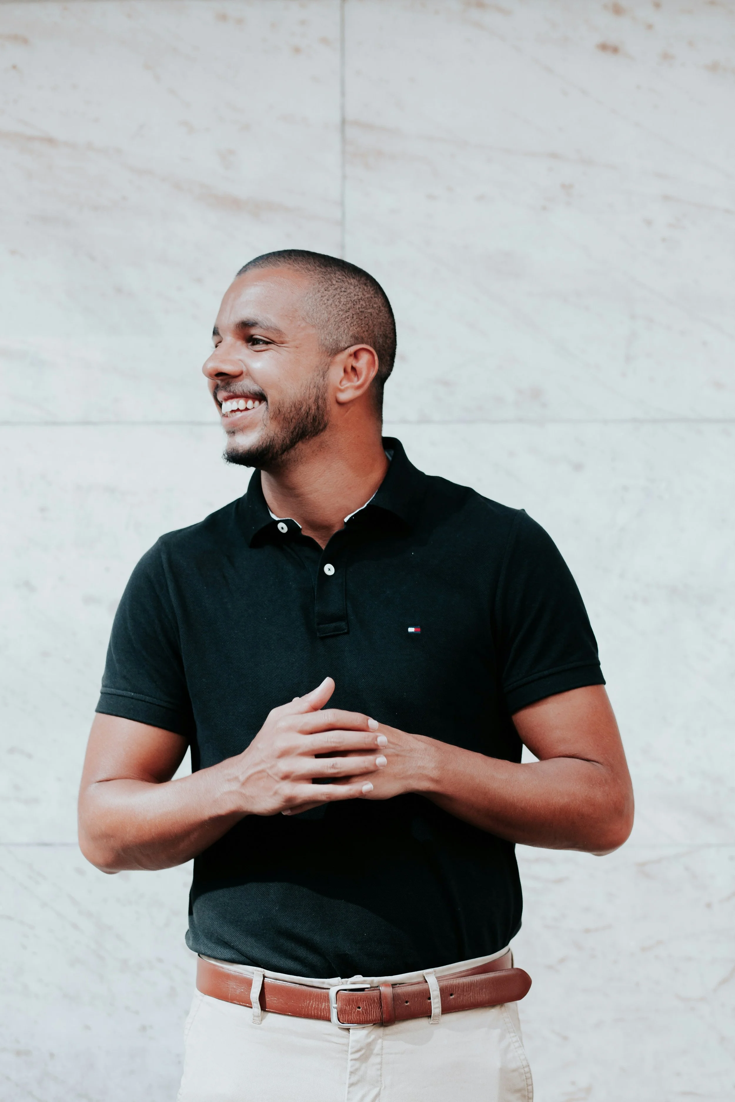 A smiling man with a trimmed beard and short hair, wearing a black polo shirt and white pants, standing against a light marble wall, appearing happy and relaxed.