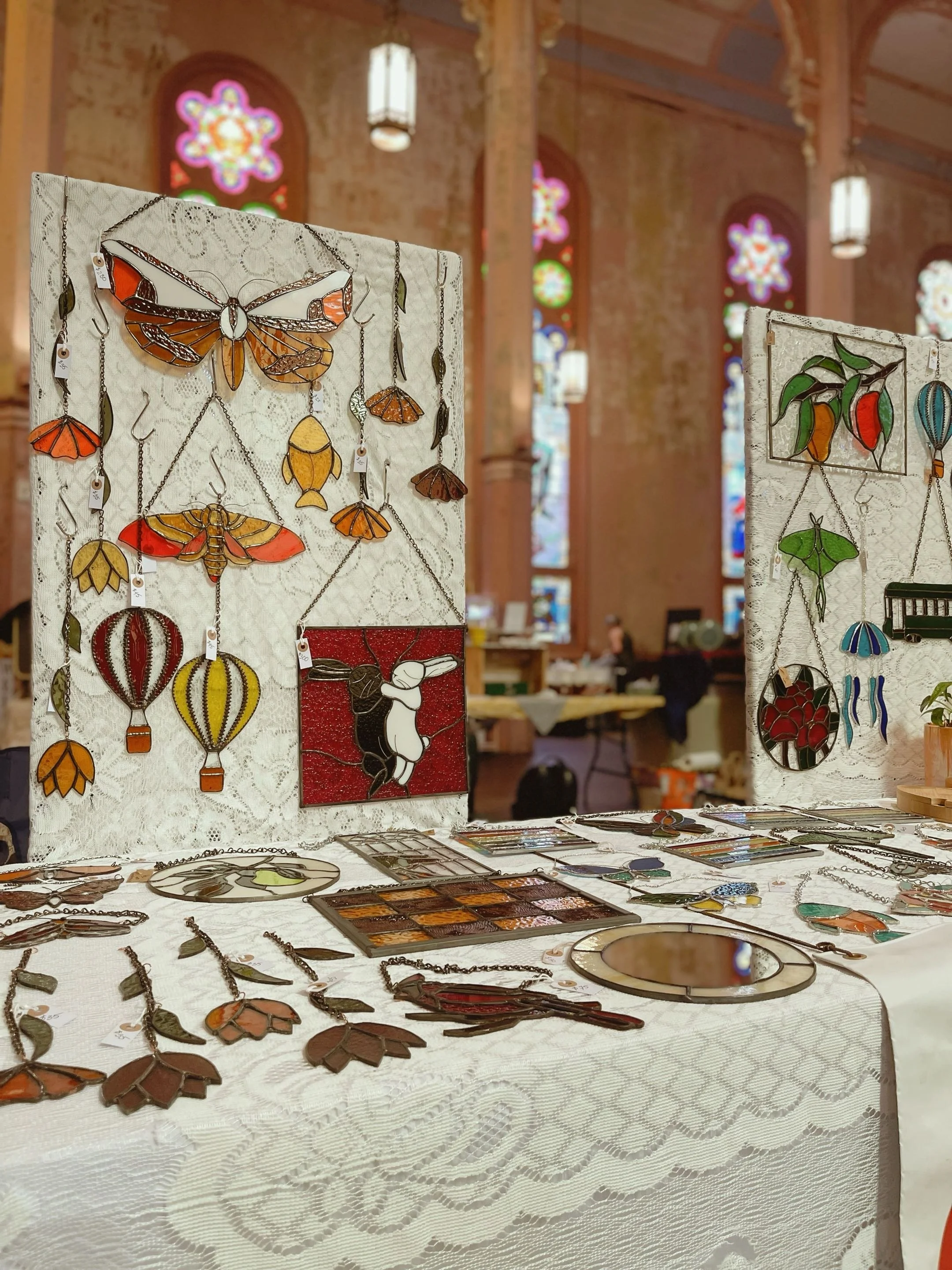 Display of stained glass artwork and jewelry at an art fair inside a building with stained glass windows and decorative lighting.