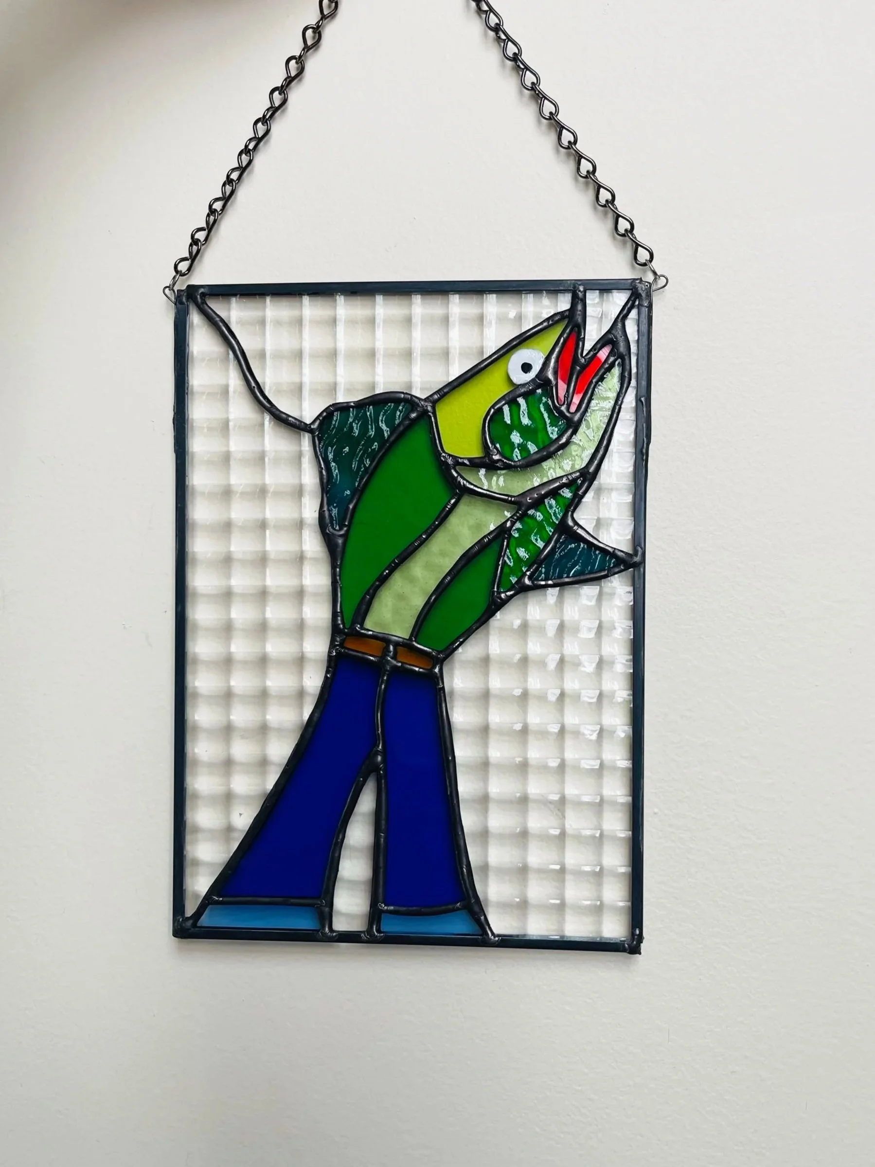 Stained glass artwork of a fish wearing bellbottom jeans, hanging on a white wall.